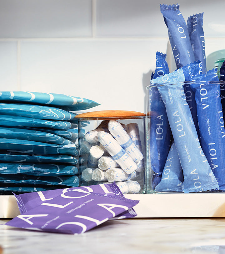 Compact plastic applicator tampons, Non-applicator tampons, Ultra thin Liners, Pads with Wings | Close-up view of a white bathroom counter with a variety of LOLA products